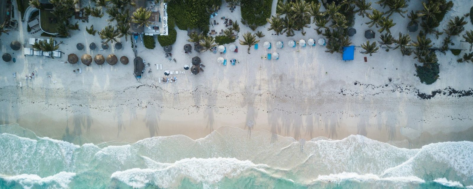 Aerial view of a Tulum beach: palm trees and beach clubs above the sand, turquoise water below
